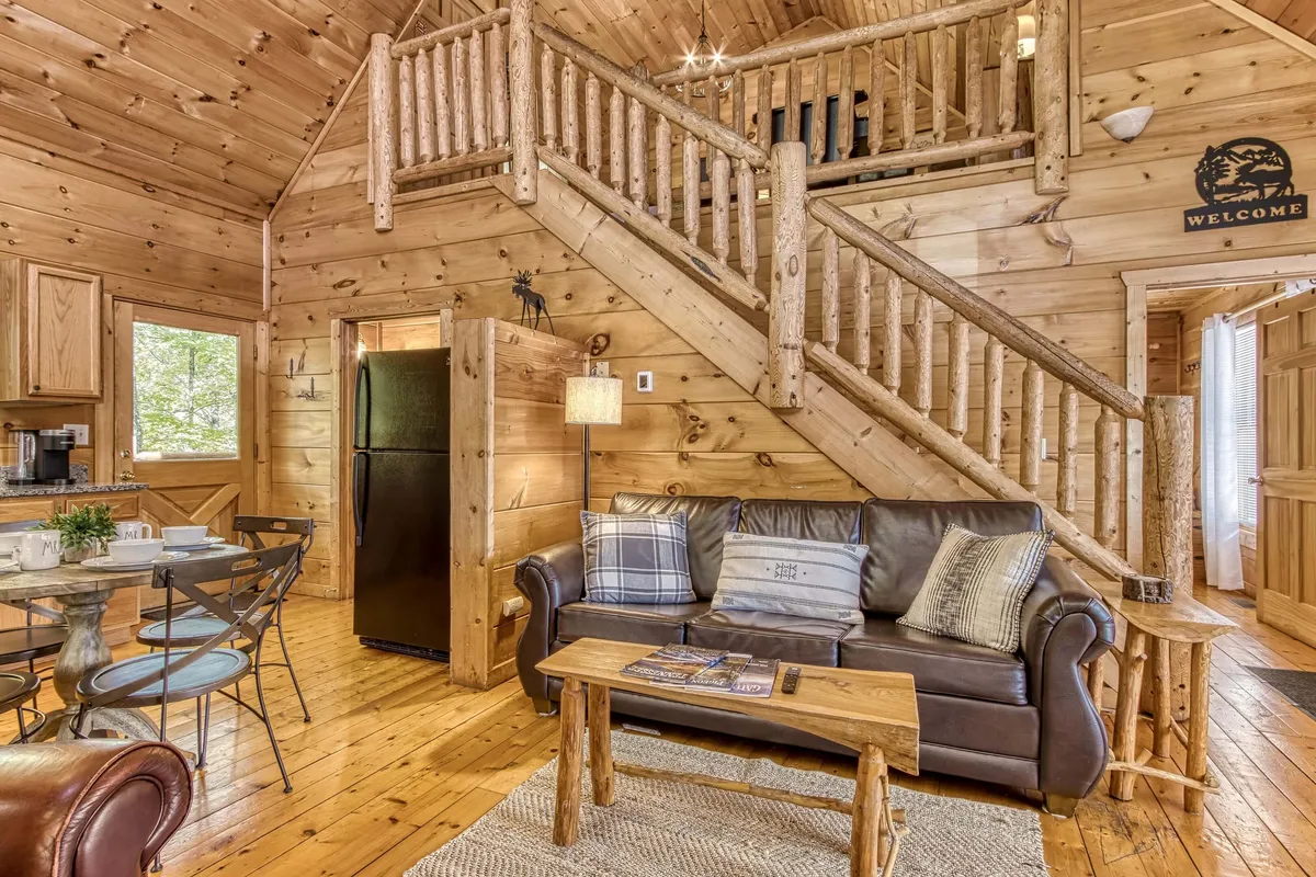 A wide view of the main living area capturing the wooden staircase, leather sofa, dining table, and kitchen area. The space is open and airy, designed for gatherings with friends or family, and displays the continuity of rustic cabin elements throughout.