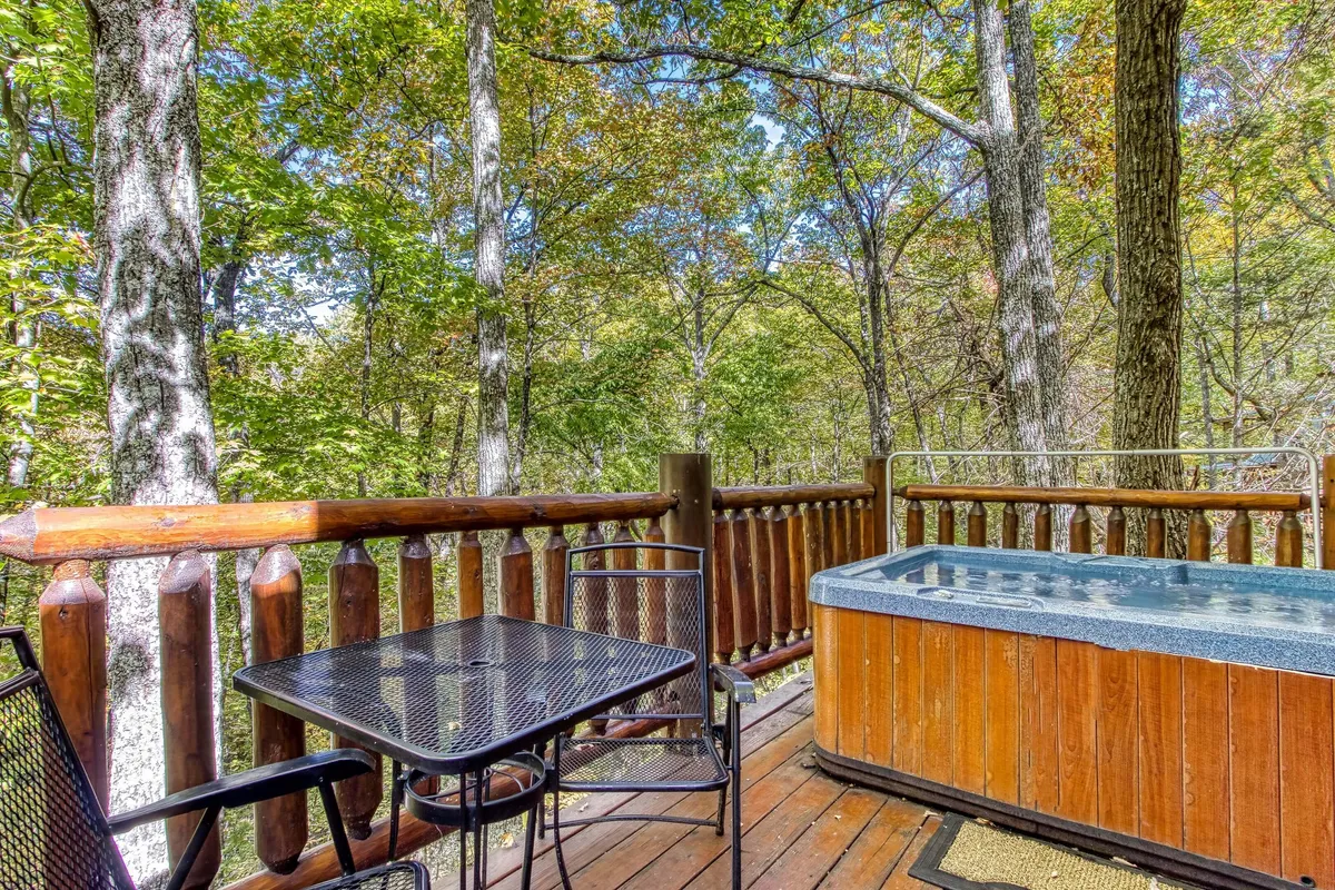 An outdoor deck with a bubbling hot tub, wooden railing, cozy table and chairs, all set amidst lush trees. The view suggests a private space perfect for soaking, with glimpses of nature and a tranquil ambiance ideal for evening relaxation or stargazing.