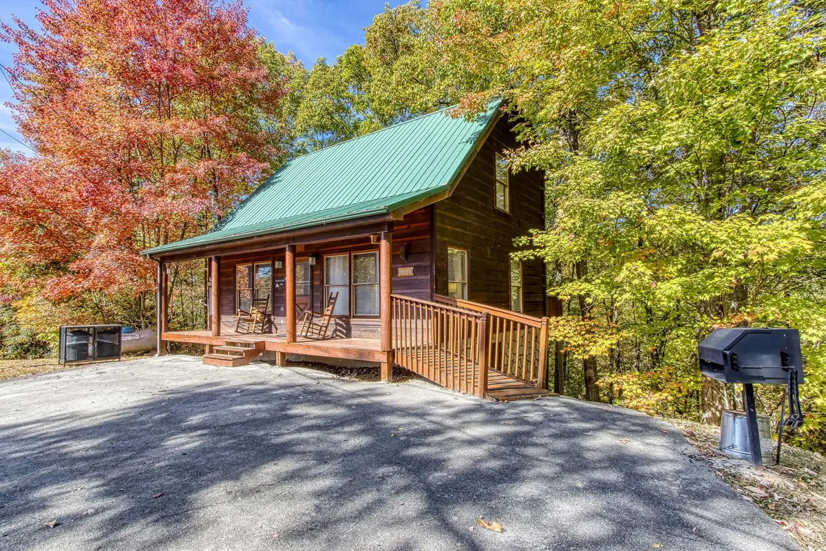 A classic log cabin exterior with a green metal roof, inviting rocking chairs on the front porch, a nearby grill, and a backdrop of vibrant autumn trees. The porch offers a welcoming space for families to relax after a day of adventure.