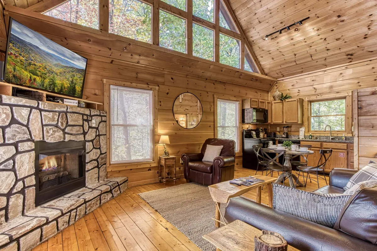 A cozy living space with a stone fireplace, leather armchair, dining table, and prominent large windows flooding the area with natural light. The blend of wood textures and inviting furnishings creates a harmonious retreat for relaxation or socializing.