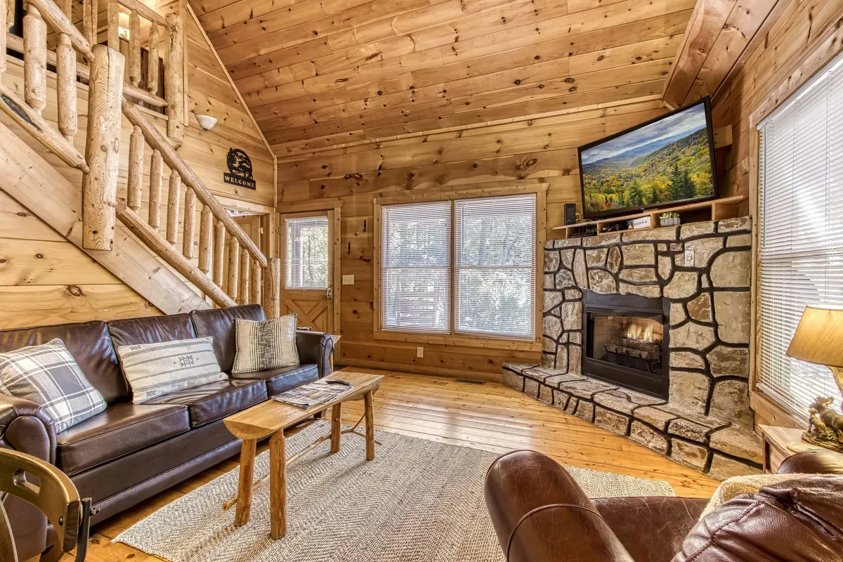 A warmly-lit living room centered on a striking stone fireplace, with a comfortable sofa, coffee table, and wooden staircase off to the side. The television is mounted above the fireplace, and the space exudes a cozy, welcoming atmosphere perfect for relaxing, watching movies, or gathering with friends and family.