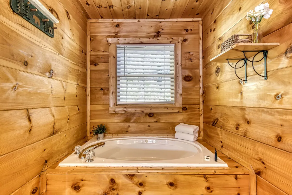 A serene indoor space featuring a deep jacuzzi tub set against wooden walls, a window with blinds admitting soft daylight, and a decorative shelf with fresh flowers. The ambiance is peaceful and inviting, ideal for unwinding after a long day.