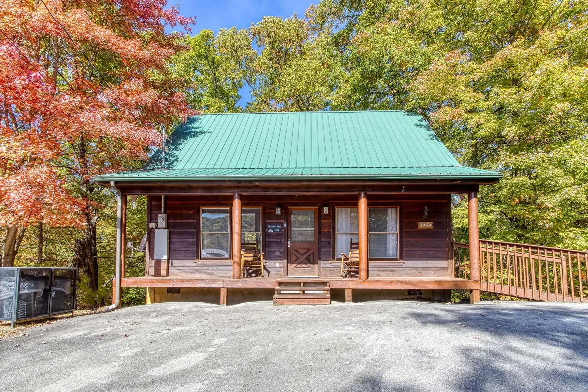 A cozy front porch with classic wooden rocking chairs overlooks colorful autumn trees, framed by a green metal roof and rustic wooden siding. The atmosphere is warm and inviting, encouraging relaxation and conversation while embracing the natural surroundings.