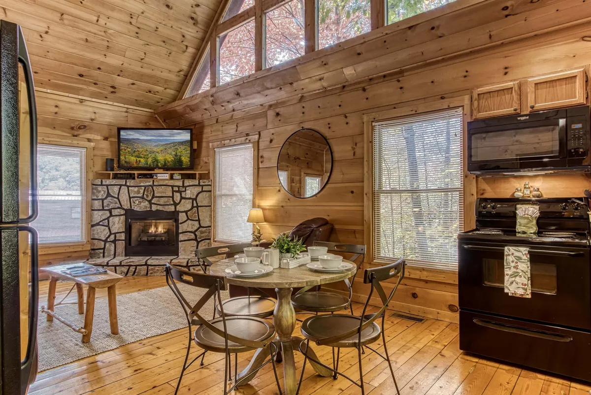 An inviting open-concept space where a large dining table and chairs sit adjacent to a stone fireplace and TV. Expansive windows fill the area with natural light, and the stone wall and wood finishes create a warm, homelike ambiance ideal for dining, entertaining, or settling in for a cozy night by the fire.