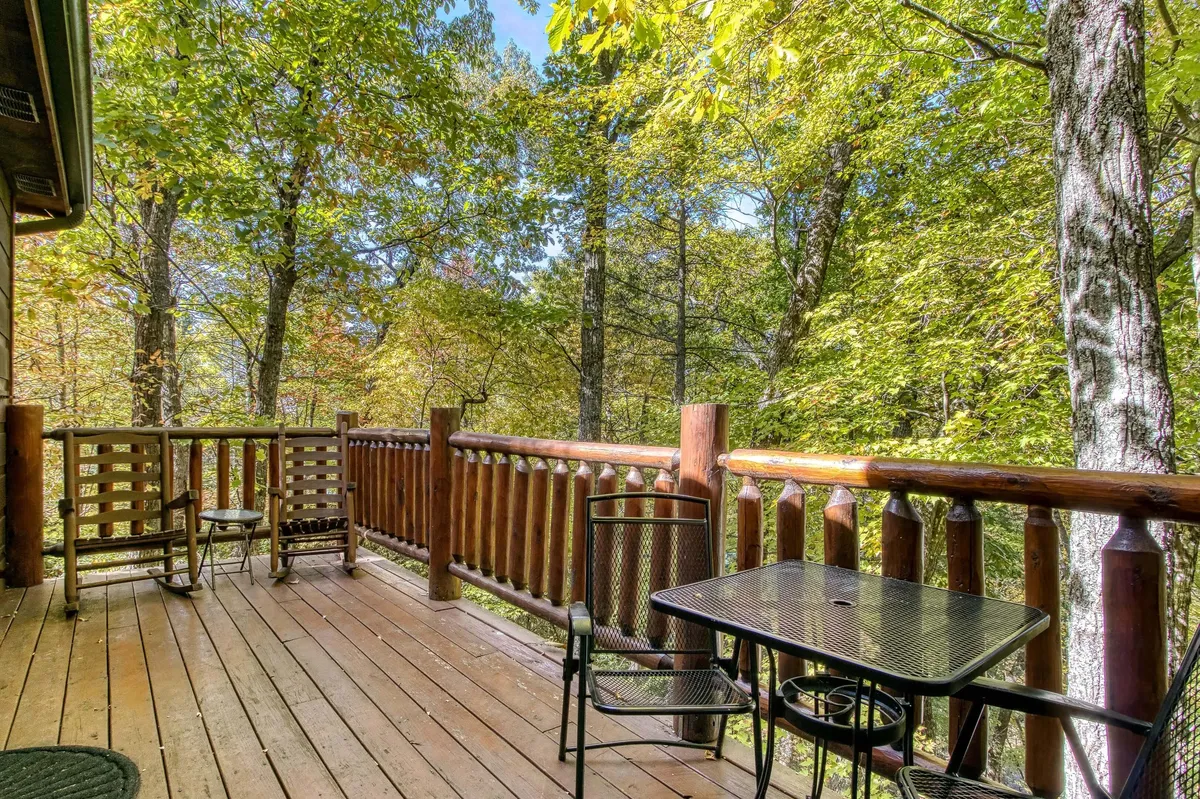 A peaceful exterior scene featuring a wooden railing, a metal table with two chairs, all surrounded by lush trees. The setup invites guests to relax while enjoying nature and the changing seasons from the comfort of the cabin’s deck.